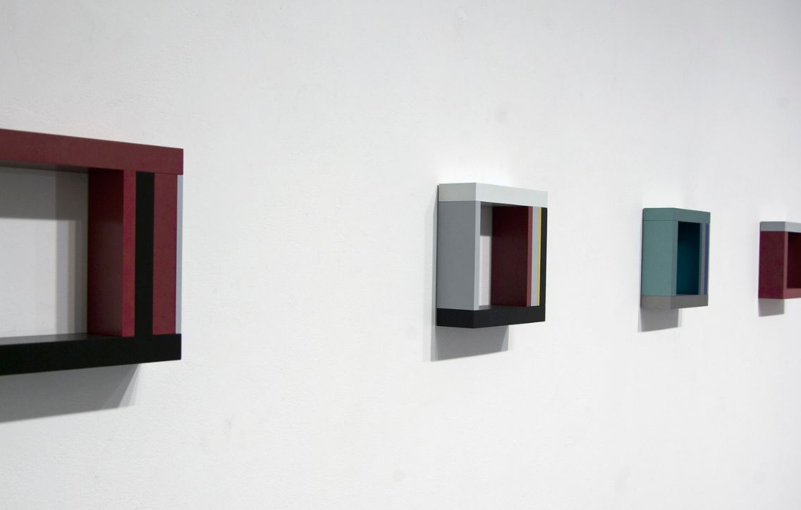 Close up image of four square sculptural art pieces mounted on a white wall in the gallery. All four pieces are painted in the same burgundy, light grey, and dark charcoal colours but each individual piece is painted with a slightly different geometric pattern.