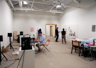 People working at the gallery. Various things are pasted o the wall, and there's also audio equipment around the place.