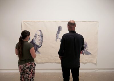 Tonya and a man looking at a piece of cloth hanging on the wall. The cloth has some faces and other things painted on it, a work in progress.