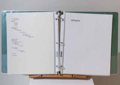 Close-up of a binder. On the left page, a poem can be seen. On the right page, there's only the word "whispers" in lowercase.